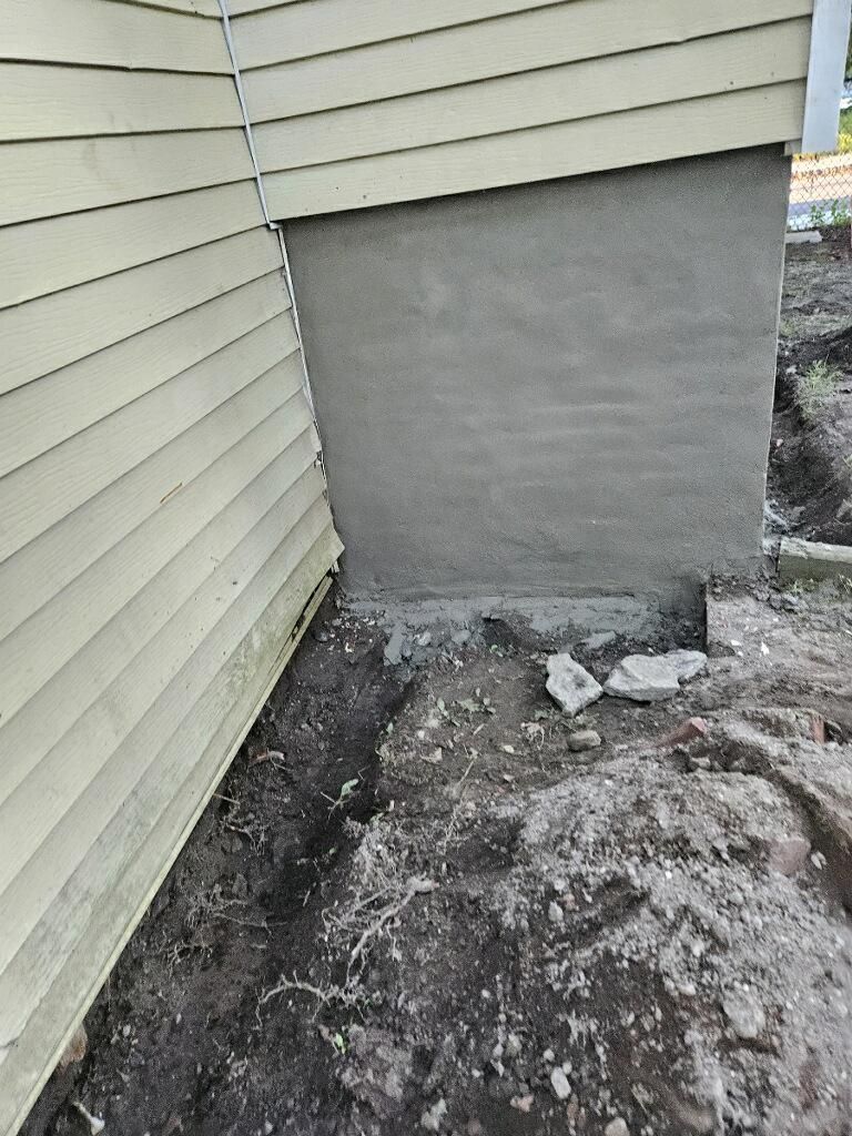 A corner of a house with a concrete wall and a hole in the ground.