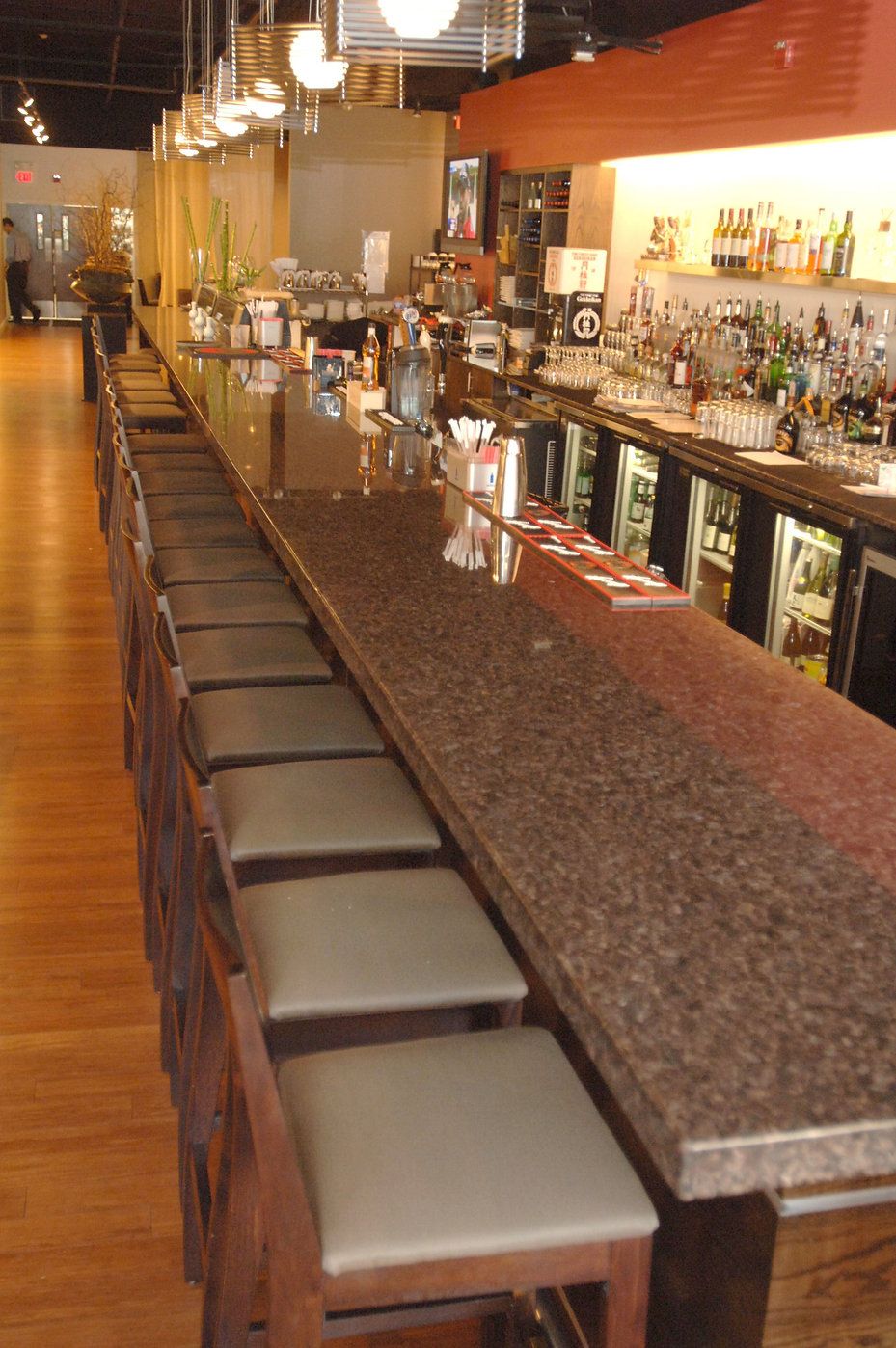 Long bar with stools in a restaurant. Granite countertop, liquor bottles, and a wooden floor.