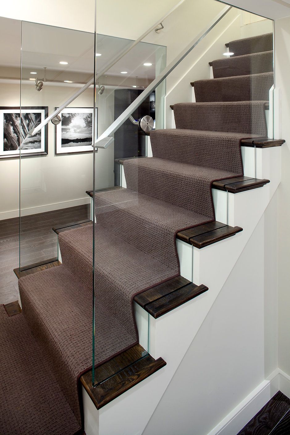 Staircase with brown carpeted steps, glass railing, and white risers, with black and white photos on the wall.