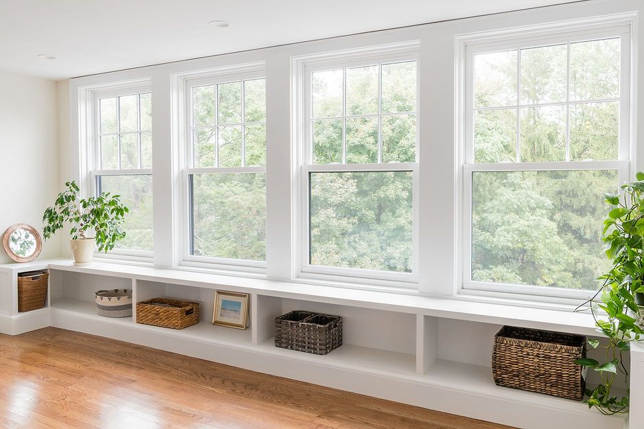 Three white framed windows overlooking trees, with a built-in shelf below, plants, and baskets.