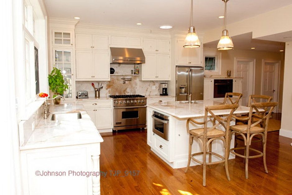 Elegant white kitchen with island and stainless steel appliances; hardwood floors and tall barstools.