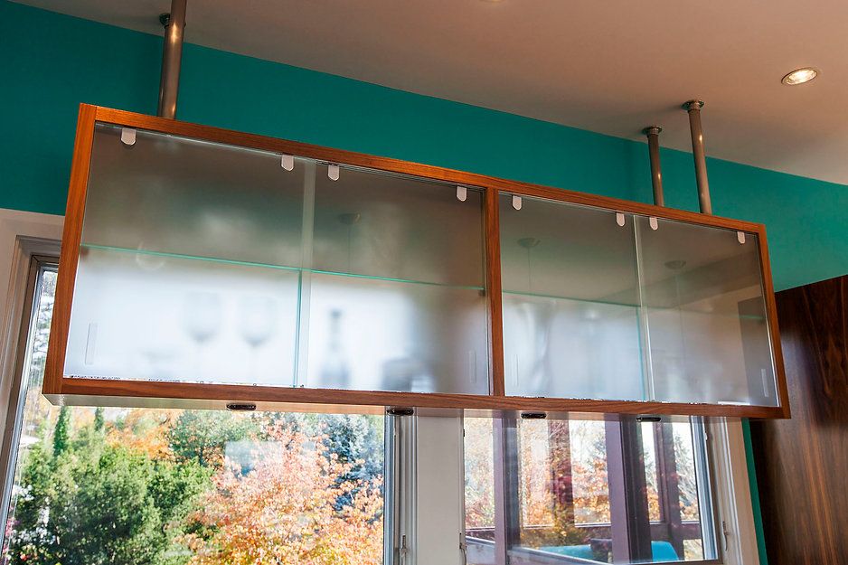 A suspended, wooden-framed glass cabinet hangs above a window, with a bright teal wall behind it.