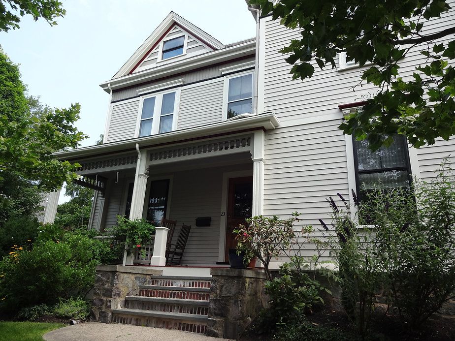 Two-story Victorian house with porch. Gray siding, stone steps, greenery. Cloudy sky.