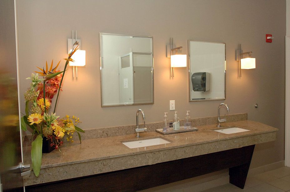 Public restroom with two sinks, mirrors, and floral arrangement, beige countertop, neutral walls.