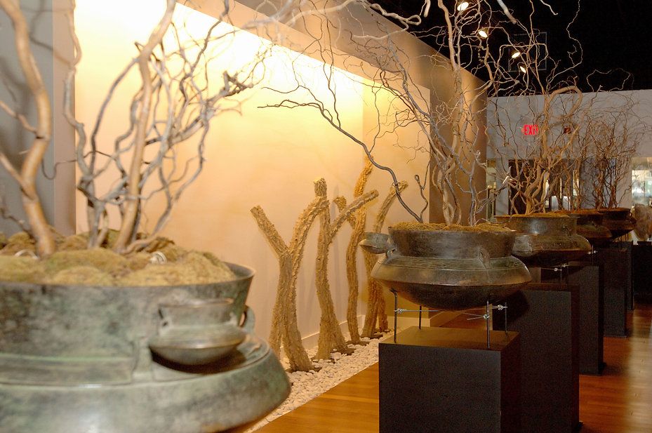 Bronze pots with bare branches on black pedestals in a gallery setting.