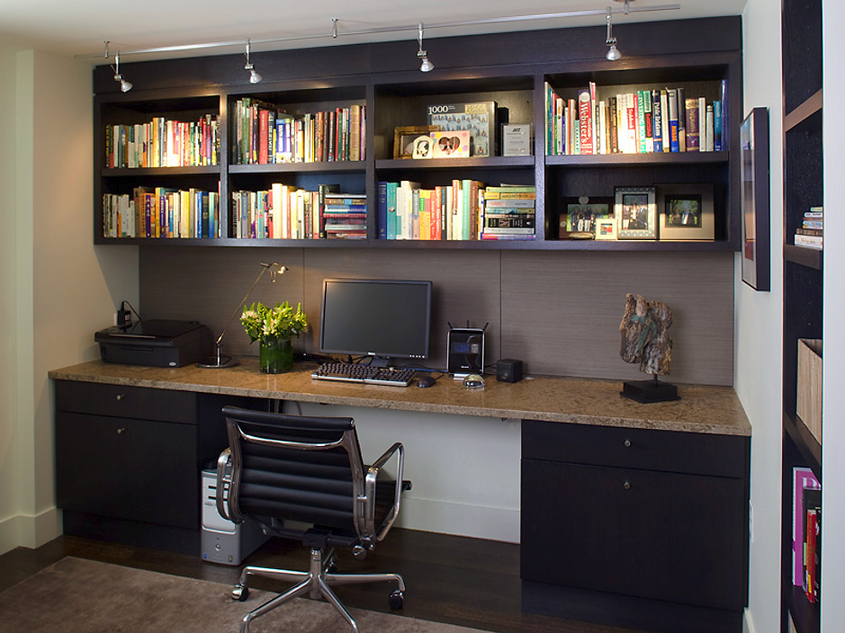 Home office with built-in desk, bookshelves, black cabinets, chair, and computer. Dark brown wood accents and beige wall.