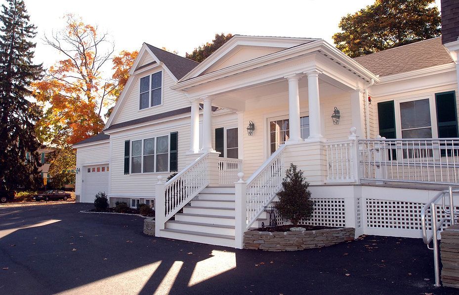 White house with porch, steps, and ramp. Driveway and trees in the background.