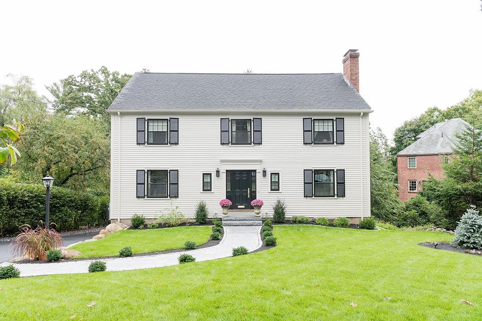 Two-story beige house with black shutters, door, and landscaping. Path leads to front door; red brick house in background.