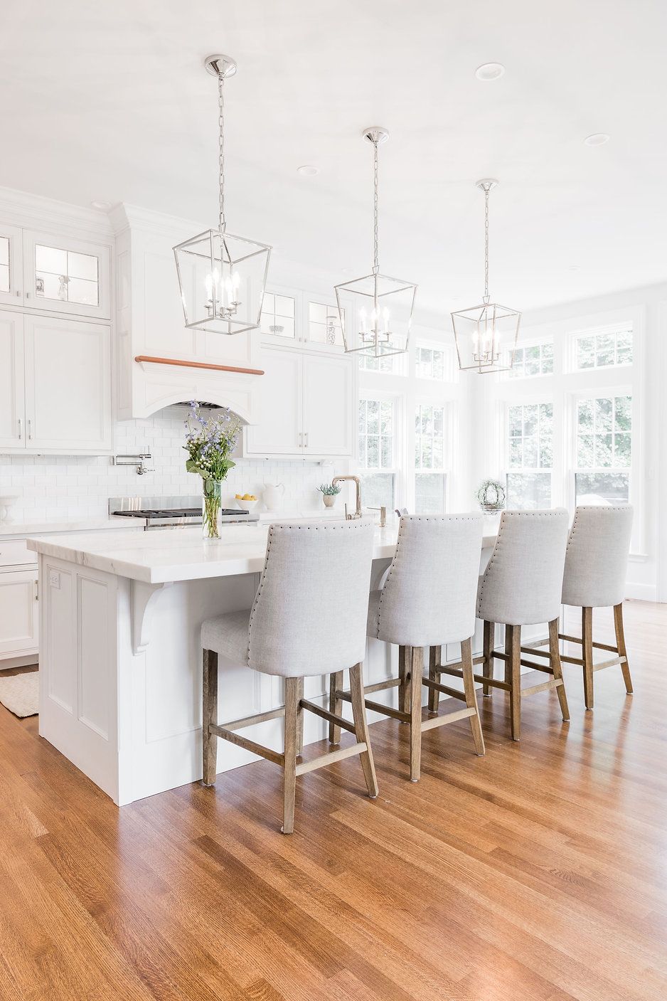 Bright, white kitchen with a center island, counter stools, and pendant lights. Wooden floors and large windows.