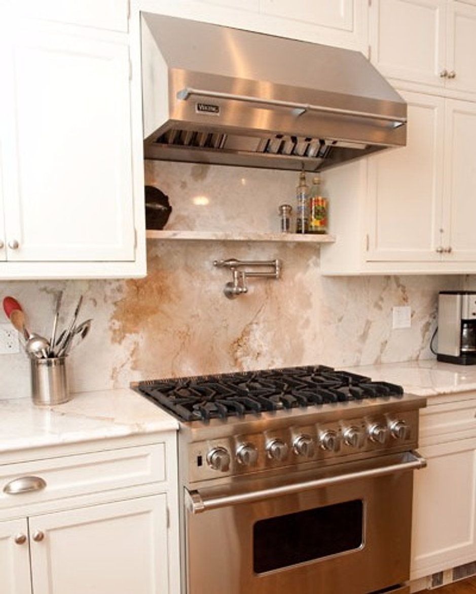 Stainless steel range hood over gas range in a white kitchen with marble backsplash.