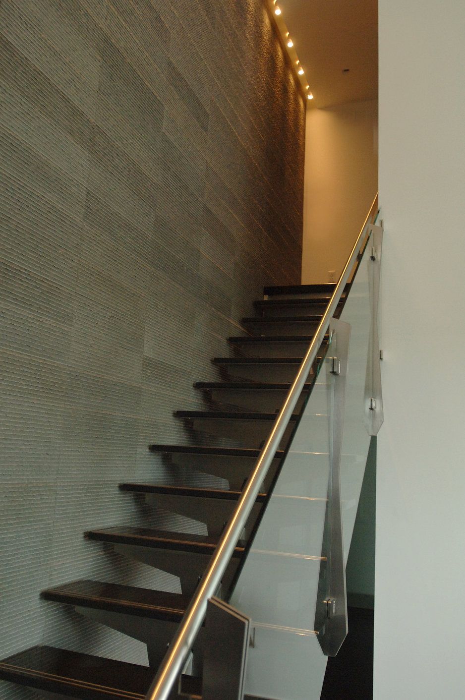 Staircase with dark wooden steps, stainless steel handrail, and textured gray wall, leading upward toward a light.