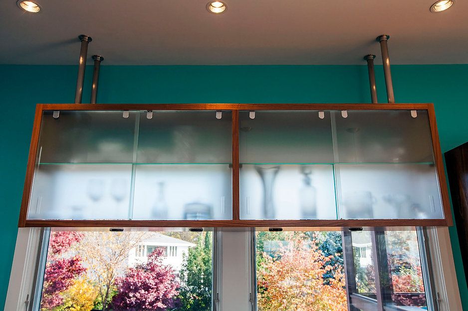 A frosted glass cabinet hangs above a window, supported by metal poles. The exterior view shows fall foliage.