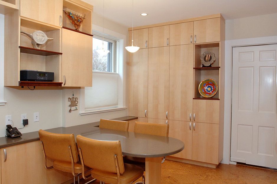 A light-wood kitchen with a round table, tan chairs, and built-in cabinets with decorative shelves.