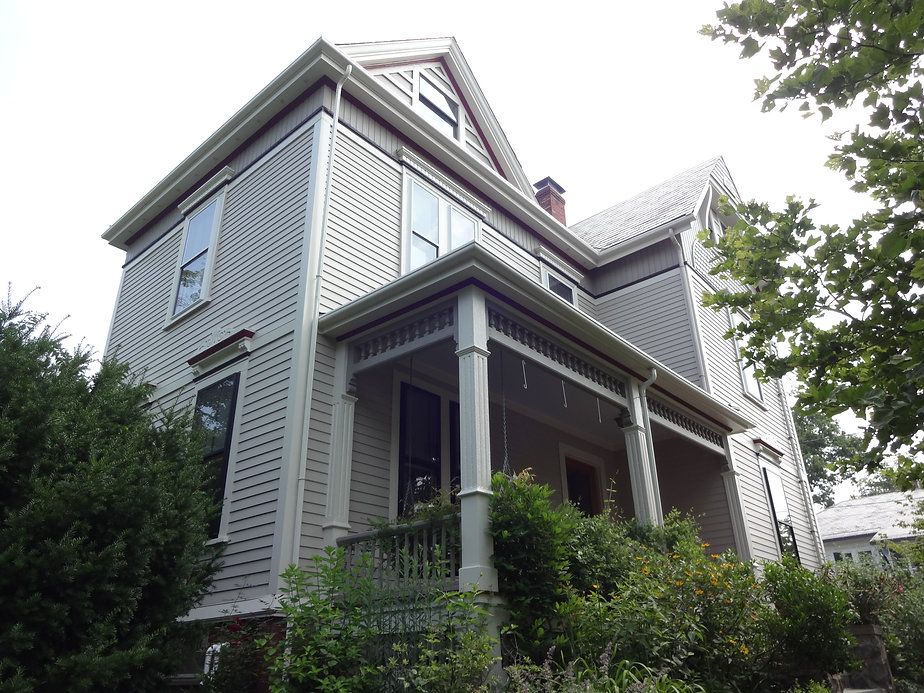 Gray Victorian house with a porch, trimmed in white, surrounded by greenery.