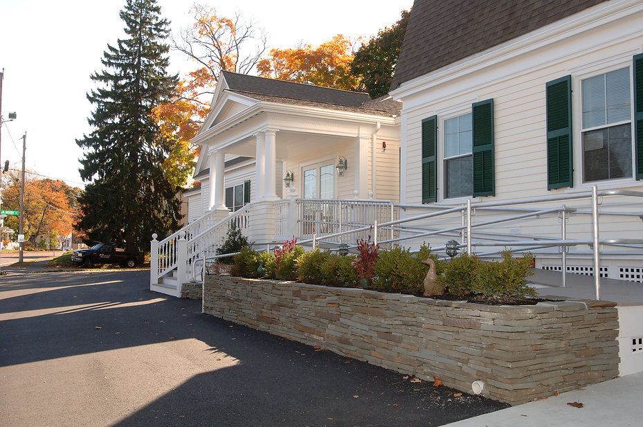Exterior view of a white building with a ramp, stone wall, and green shrubs.