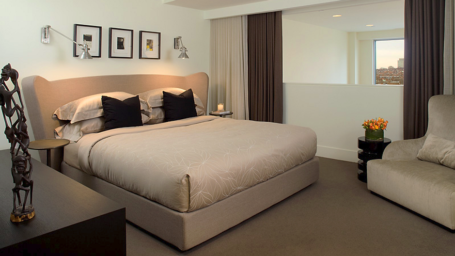 Modern bedroom with a large bed, black pillows, and an armchair. Neutral colors with artwork on the wall.