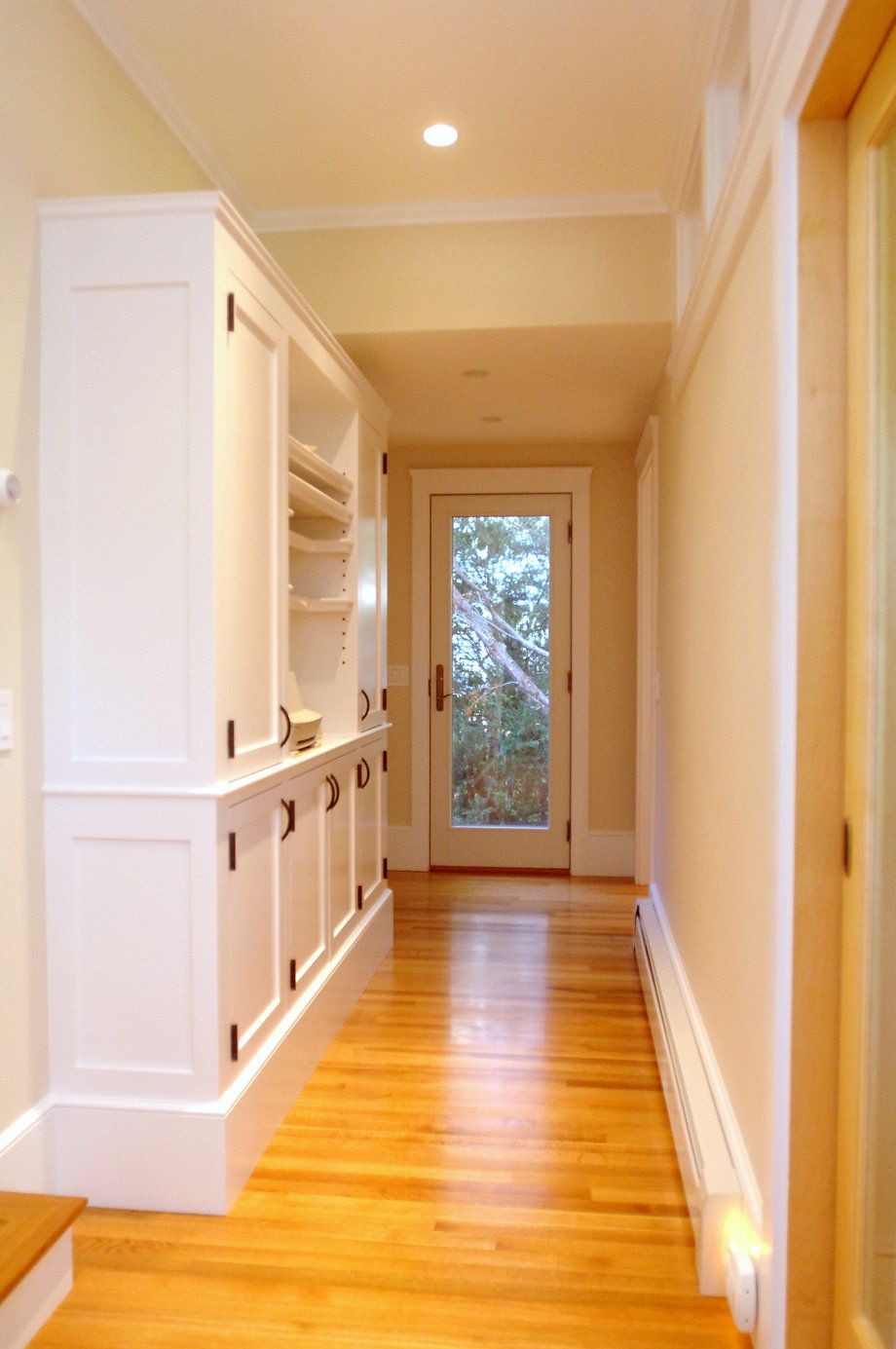 Hallway with white built-in cabinets on one side, wooden floor, and door at the end looking outside.