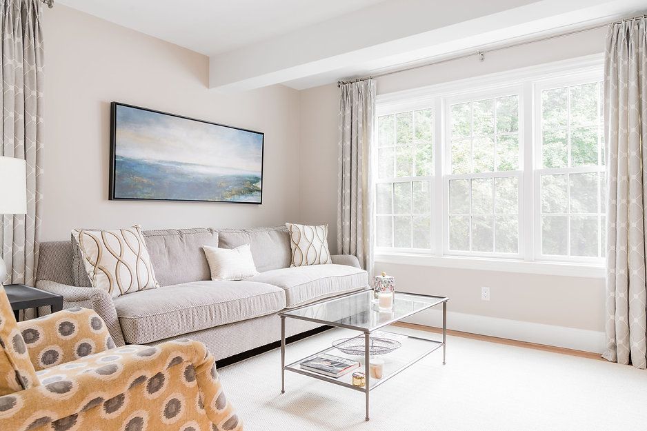 Cozy living room with gray sofa, patterned armchair, and large window with white trim and curtains.