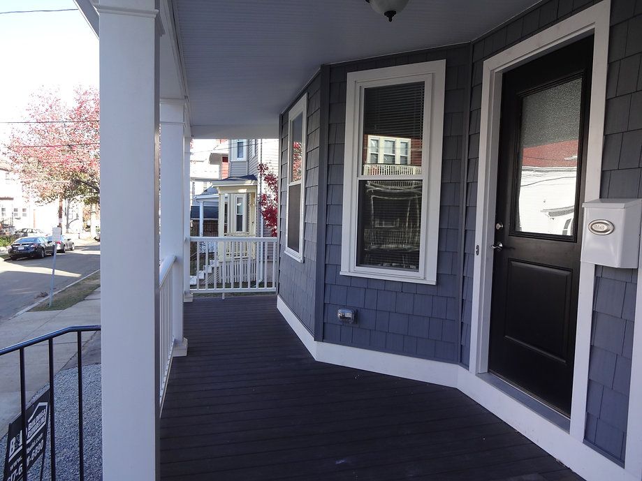 Front porch of a house with blue siding, black door, and white railing, overlooking a street.