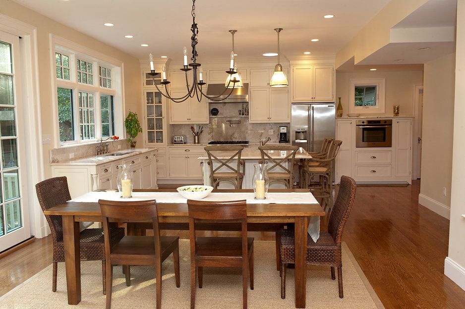 Dining area with wooden table and chairs, open to a kitchen. Light-filled room with neutral tones and modern appliances.