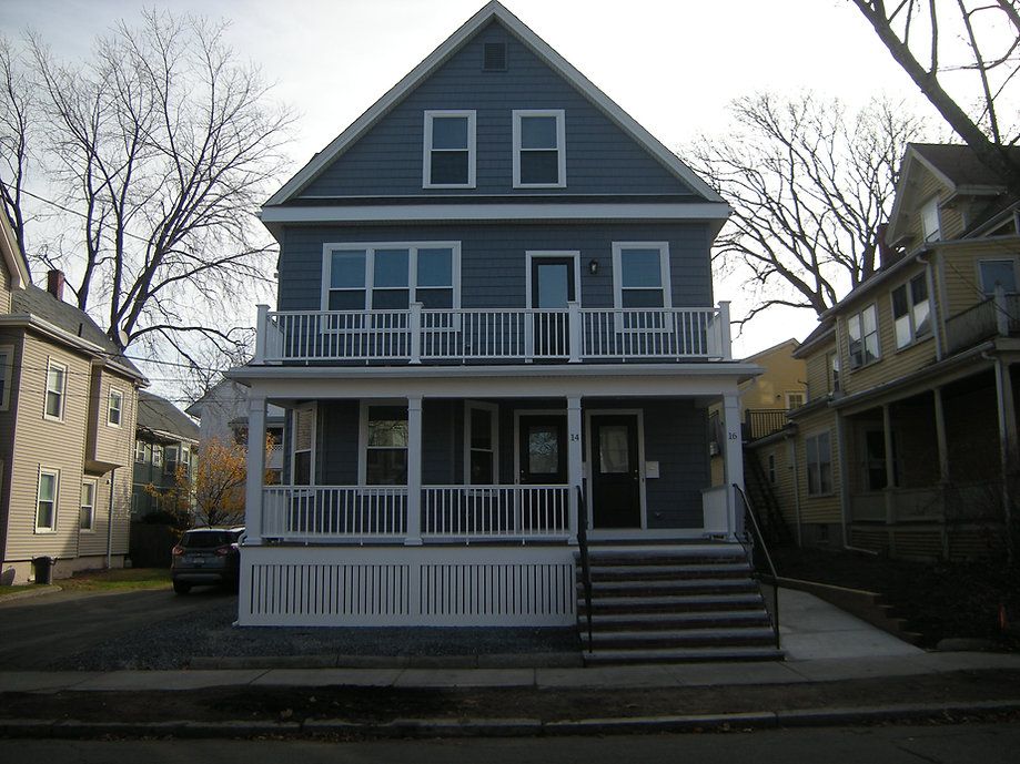 Two-story blue house with white railings and steps leading to the front door, flanked by other houses on a city street.