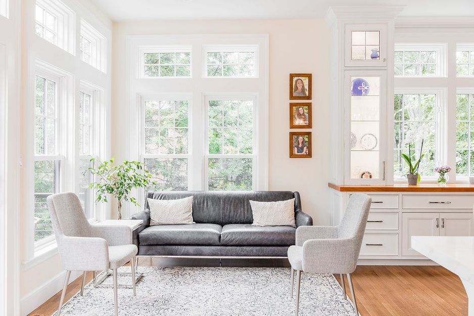 A light-filled living room with a gray sofa, two armchairs, and large windows overlooking trees.