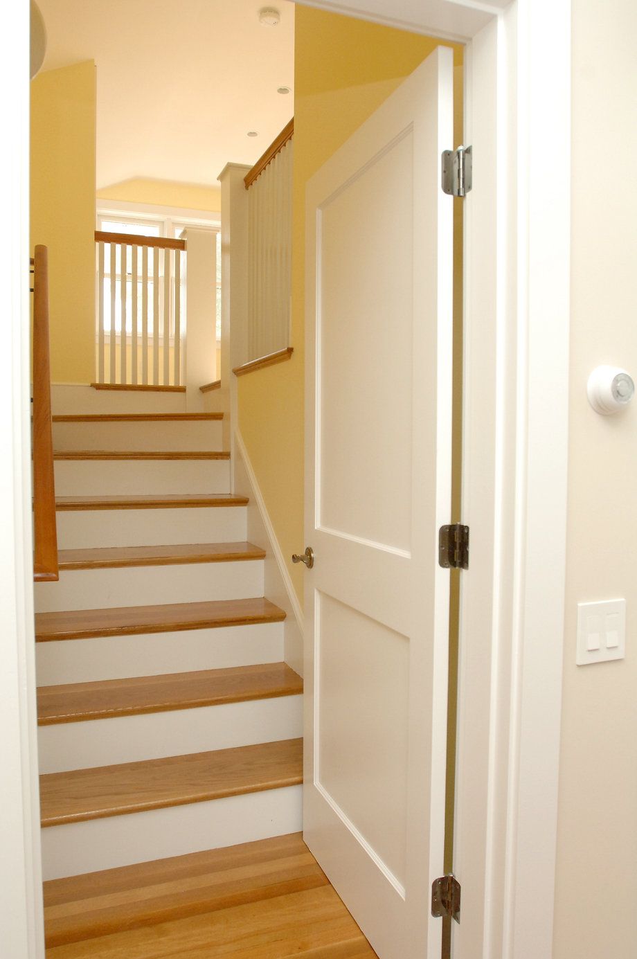 Open white door reveals staircase with wooden steps and railings. Yellow walls and light-colored trim.