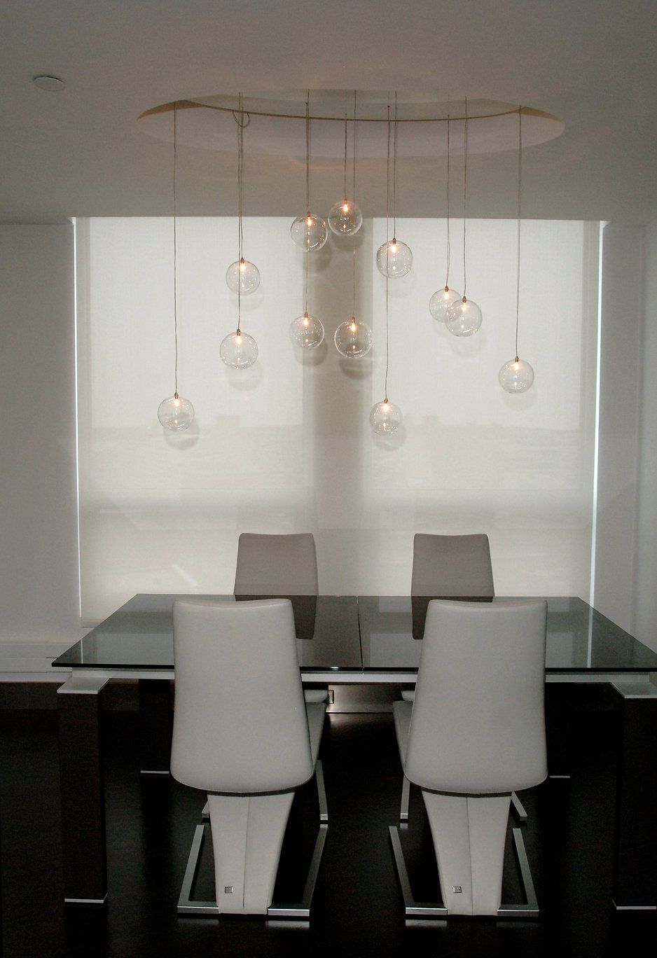 Dining room with a glass table, white chairs, and pendant lights over a window with blinds.