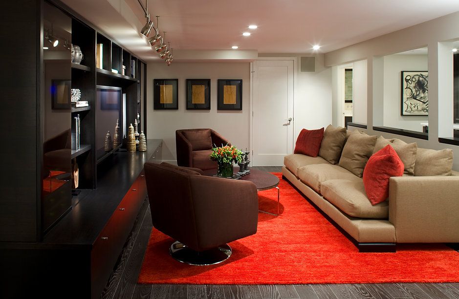Modern living room with red rug, tan sofa, brown chairs, dark shelving.