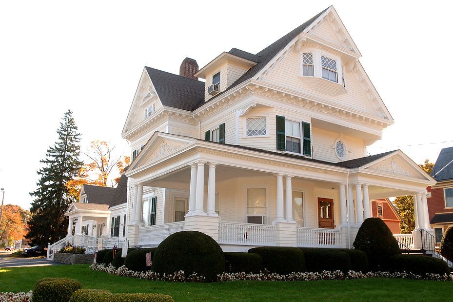 White Victorian house with porch and columns, on a lawn, with trees and sky.