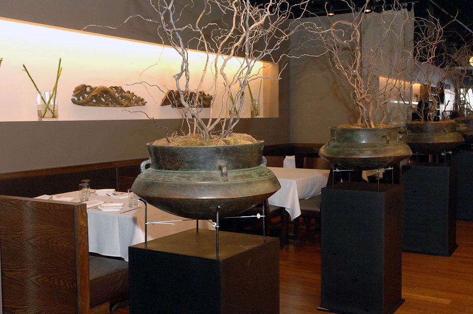Restaurant interior with antique urns on pedestals holding bare branches, tables set for dining.