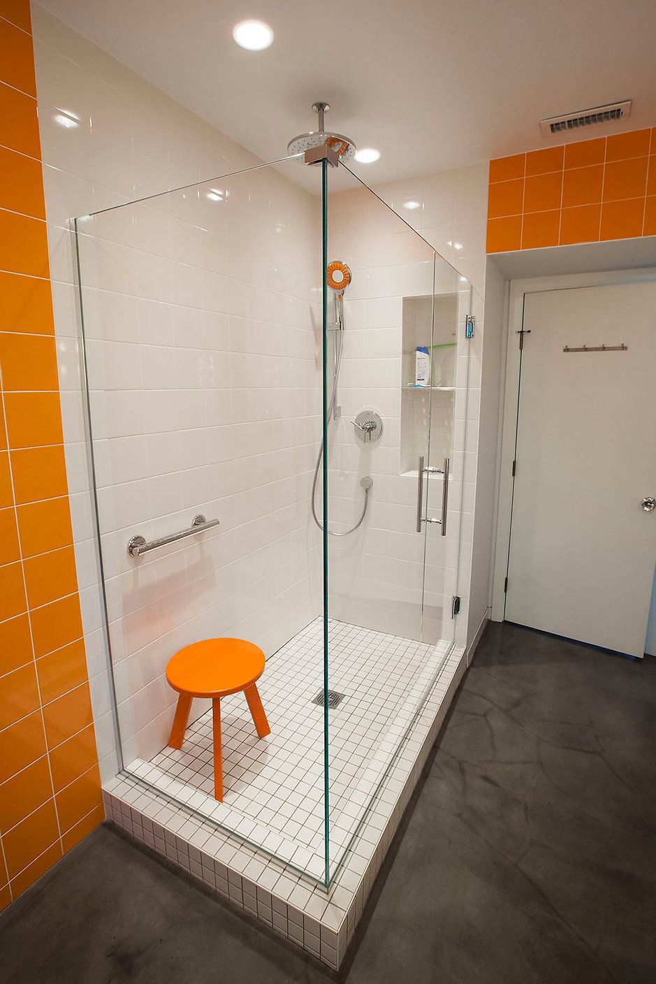 Bright orange and white tiled shower with glass door, small orange stool, and grab bar.