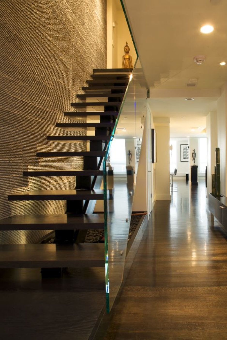 Modern staircase with floating dark wood steps, glass railing, and Buddha statue. Hallway with wood flooring.