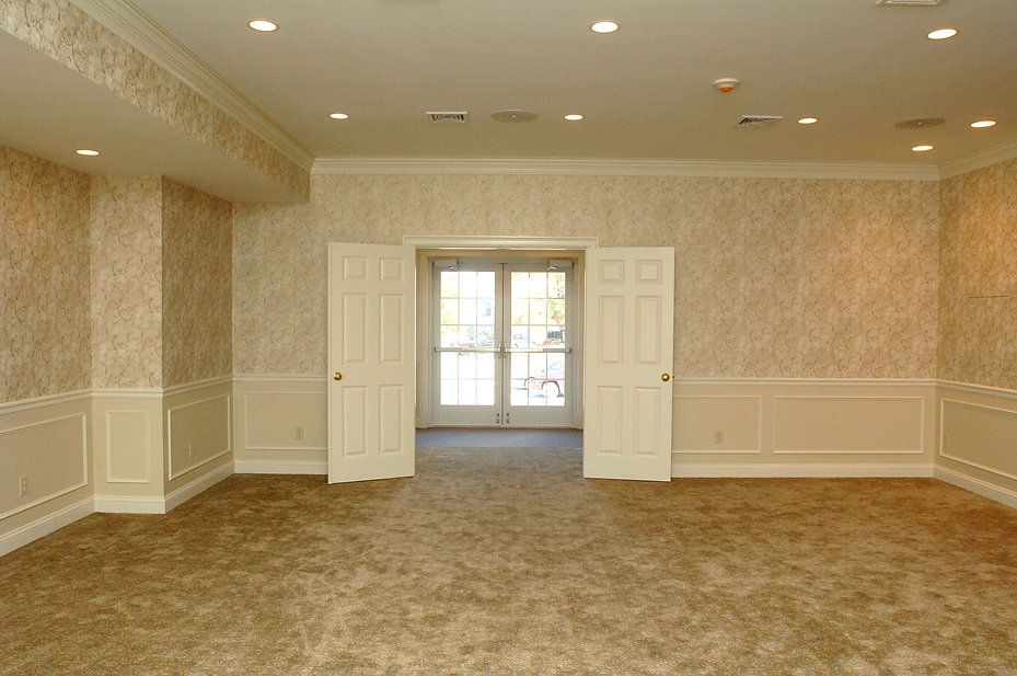 Empty room with beige patterned wallpaper, carpet, and wainscoting. French doors open to the outside.