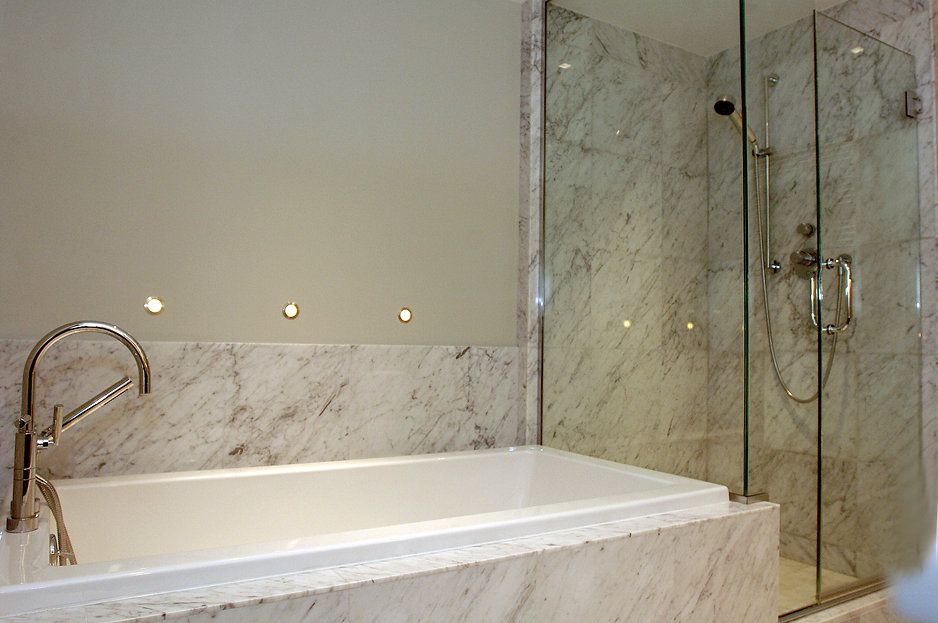 Bathtub and shower enclosed by glass. Marble walls and trim. Chrome faucet and fixtures.