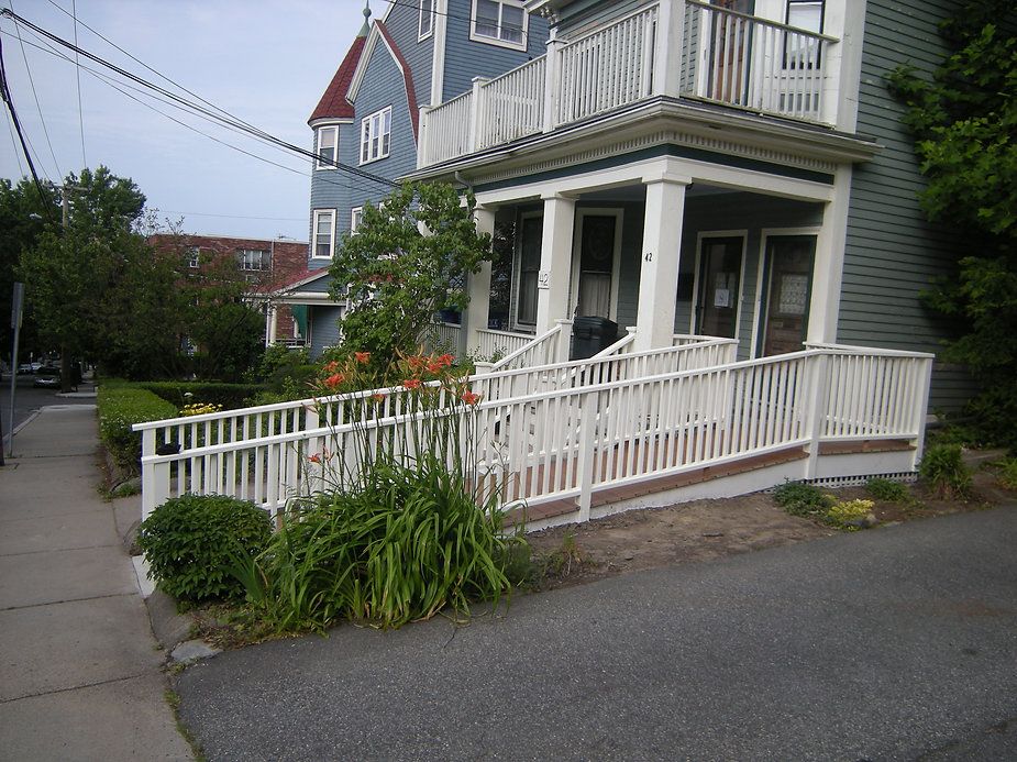 A light green house with a white ramp, sidewalk, and garden. Other houses and the street are visible.