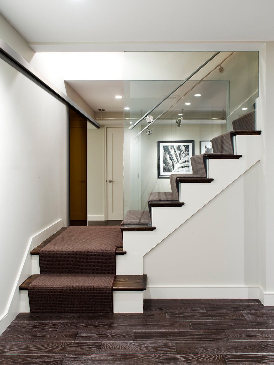 Staircase with carpeted steps, glass railing, white walls, and dark wood flooring.