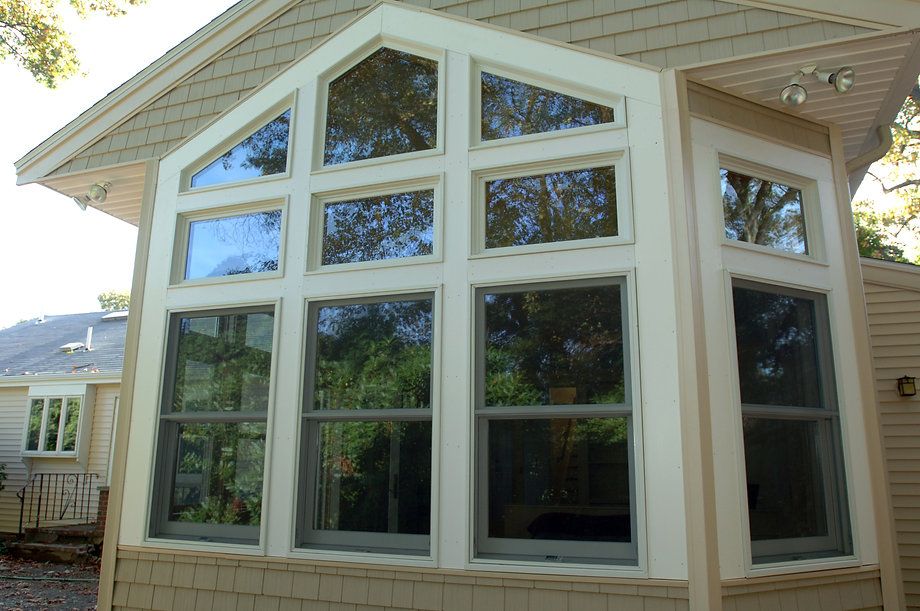 Exterior view of a house with large windows. White trim, beige siding. Trees visible through the glass.