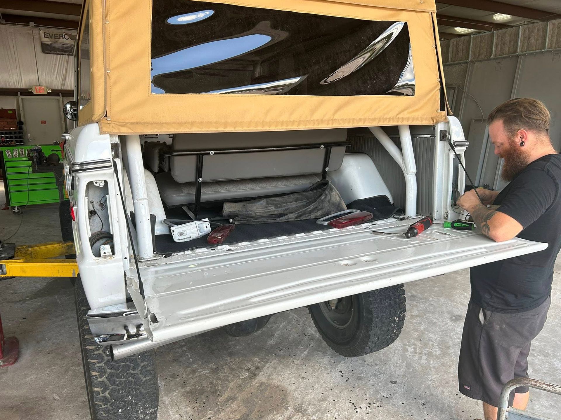 Man working on the open back of a white vintage SUV in a garage, tan top, tools present.