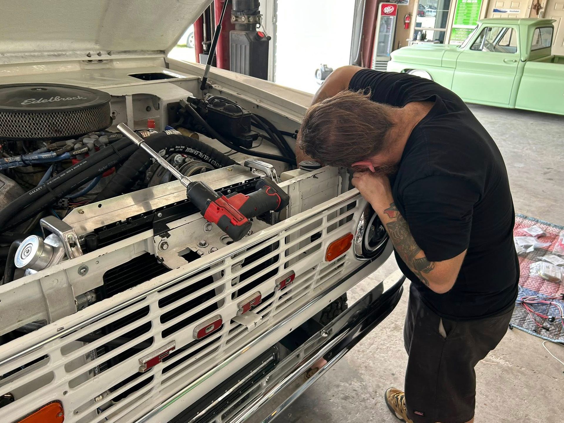 Man working on a white vintage Bronco's engine in a garage, using a power tool.