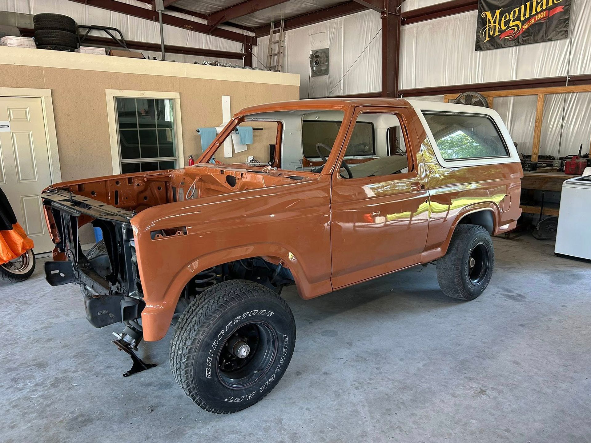 A vintage brown Ford Bronco undergoing restoration in a garage. The vehicle is partially disassembled, with black wheels.