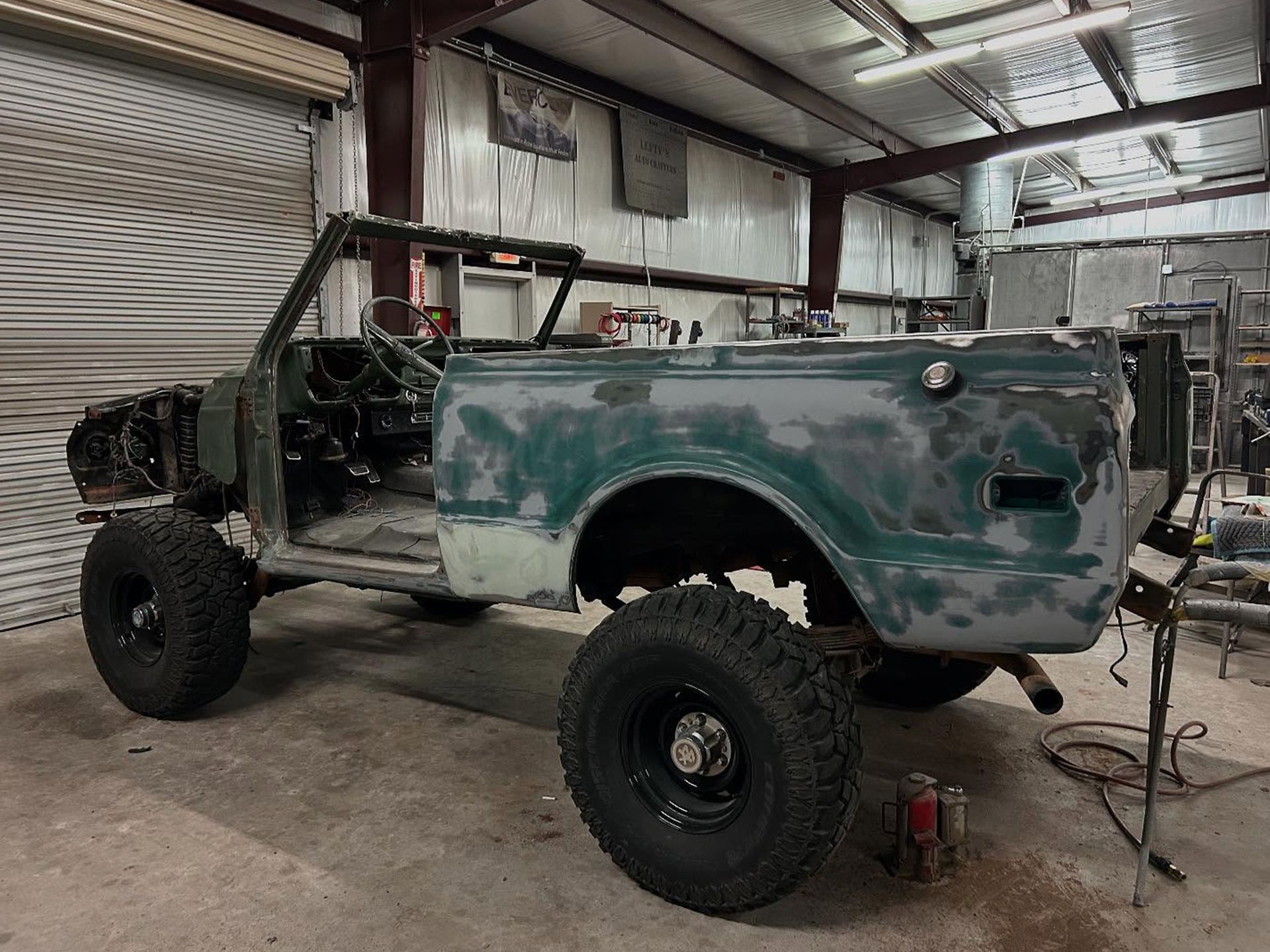 Green vintage SUV undergoing restoration in a garage, with exposed frame, large tires, and body panels.