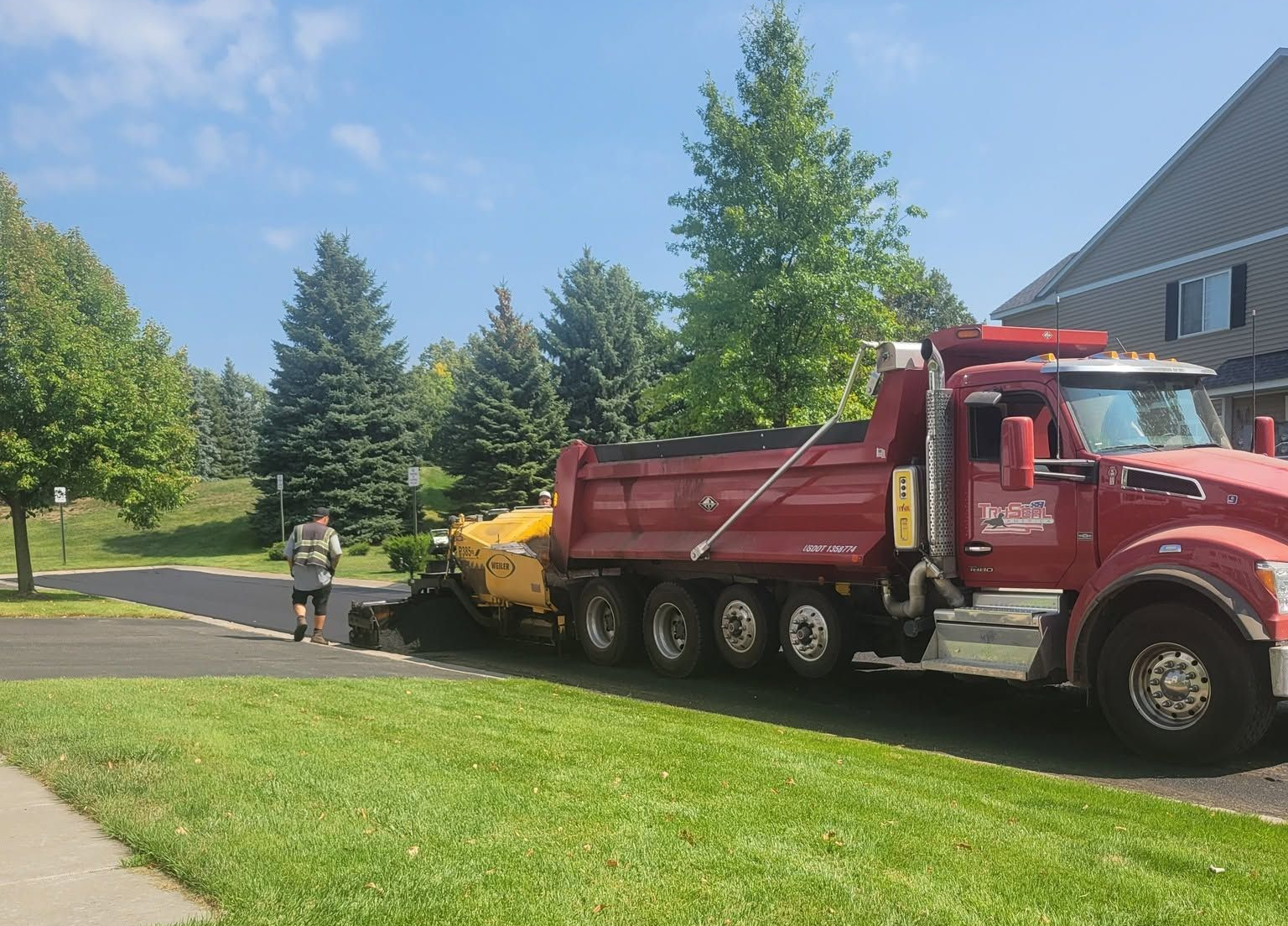 Red dump truck and worker beside yellow equipment on a sunny suburban street with trees and grass.