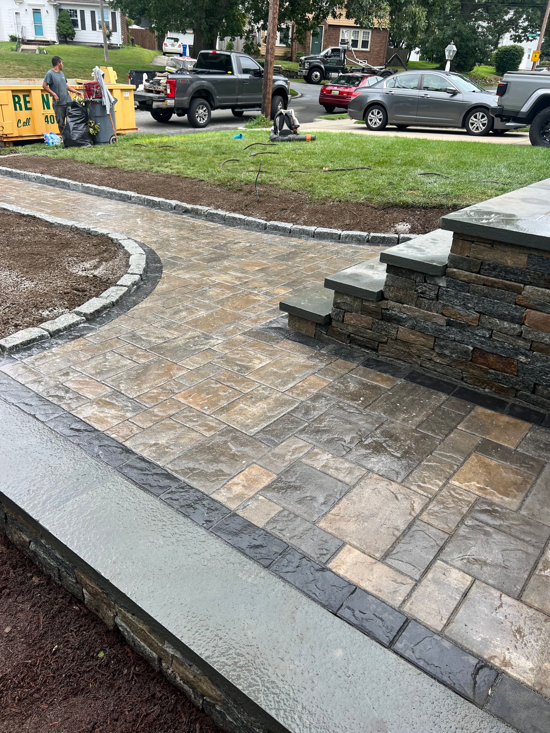 A brick walkway is being built in a park with cars parked on the side of the road.
