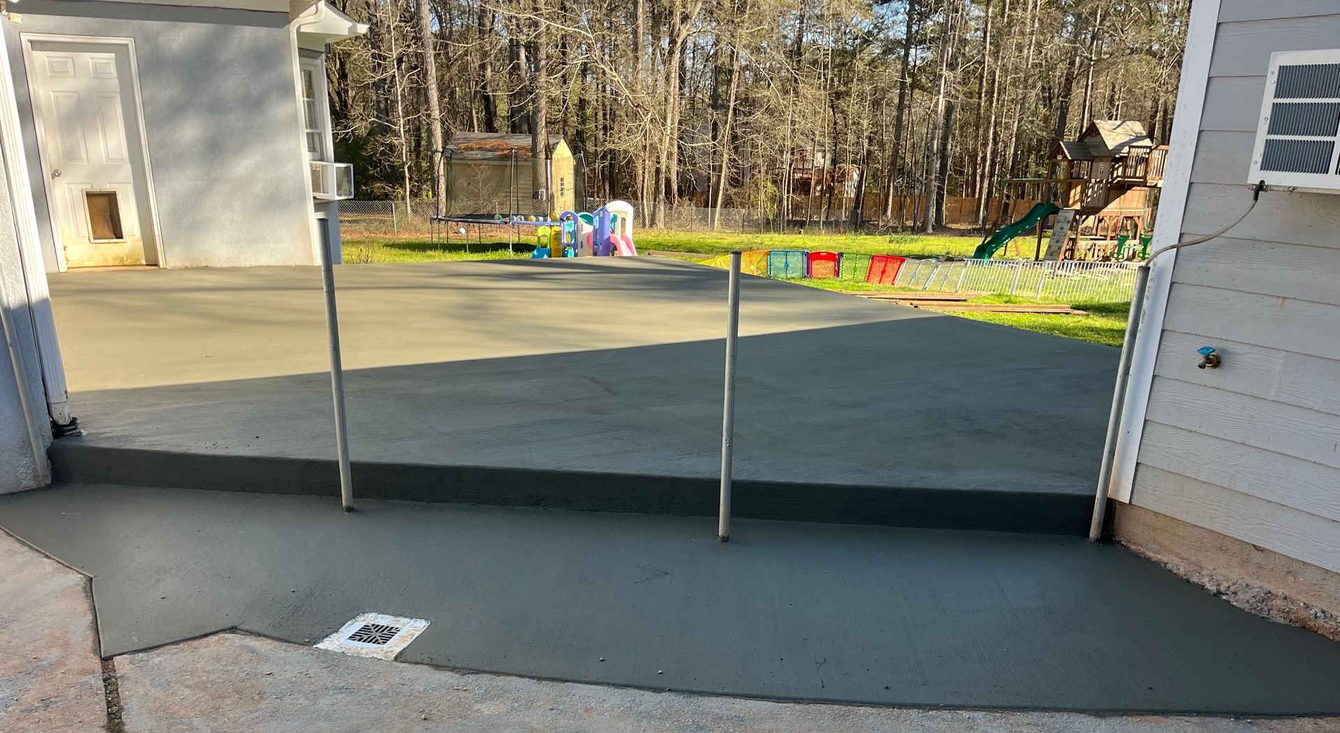 A freshly poured gray concrete patio extension featuring a step down, a drainage grate, and a metal fence post.