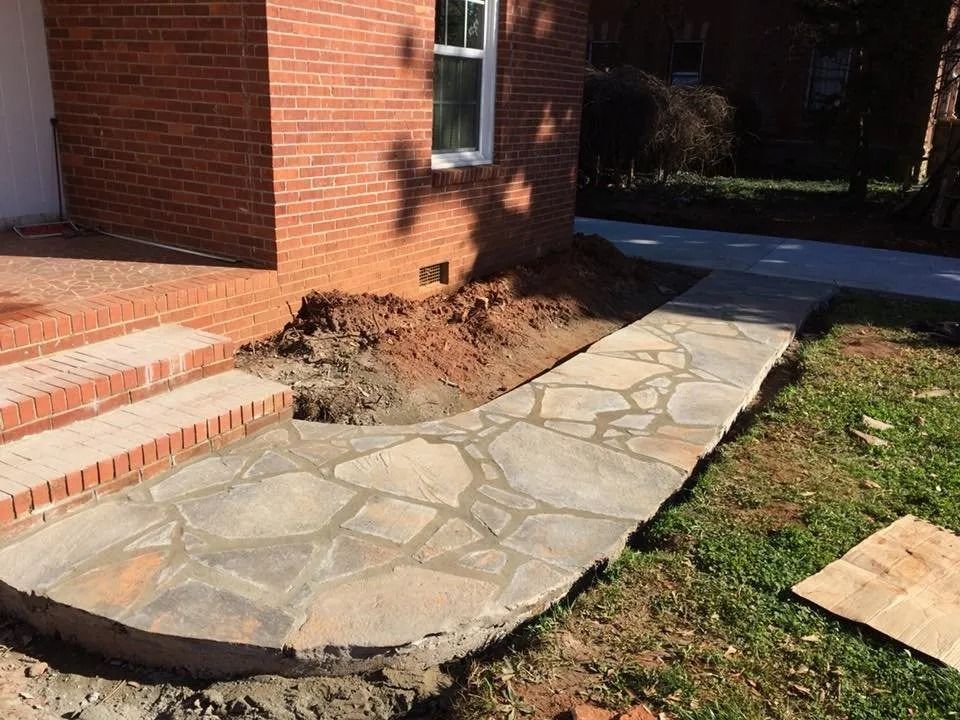 A newly installed flagstone walkway leads to brick front steps of a house, with fresh dirt along the building's edge.