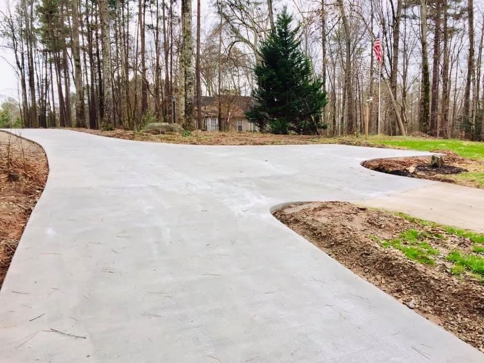 A newly poured gray concrete driveway splits into two paths, surrounded by a wooded yard with scattered mulch.