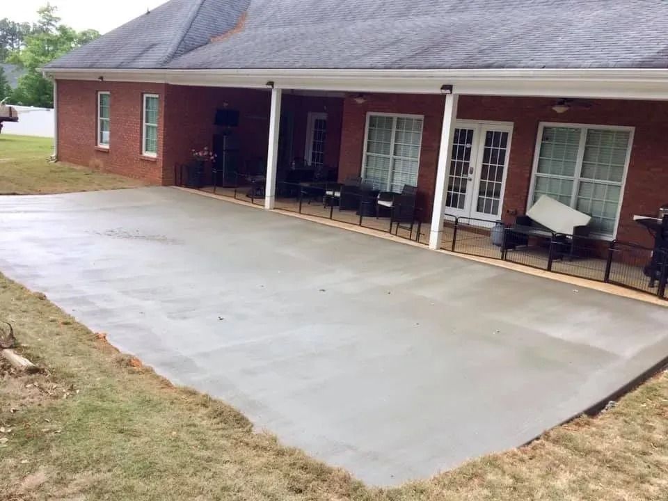 Newly poured concrete patio extending from a brick house with a covered porch and patio furniture.
