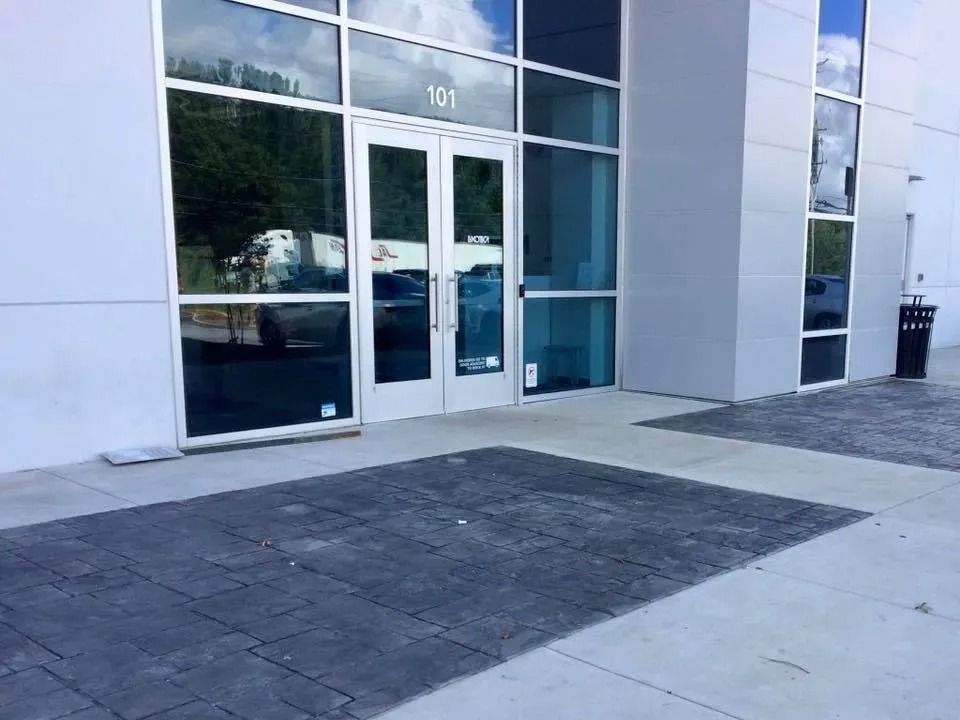 A modern building exterior entrance with glass double doors numbered 101, flanked by large windows and a paved walkway.