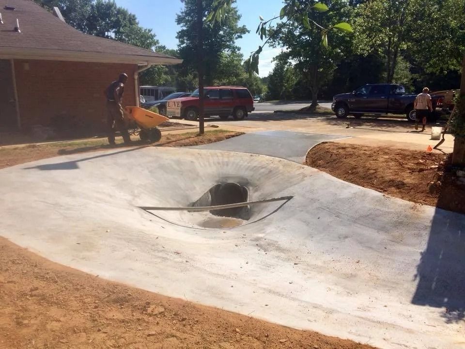 A construction worker uses a wheelbarrow near a newly installed concrete drainage basin in a residential area.